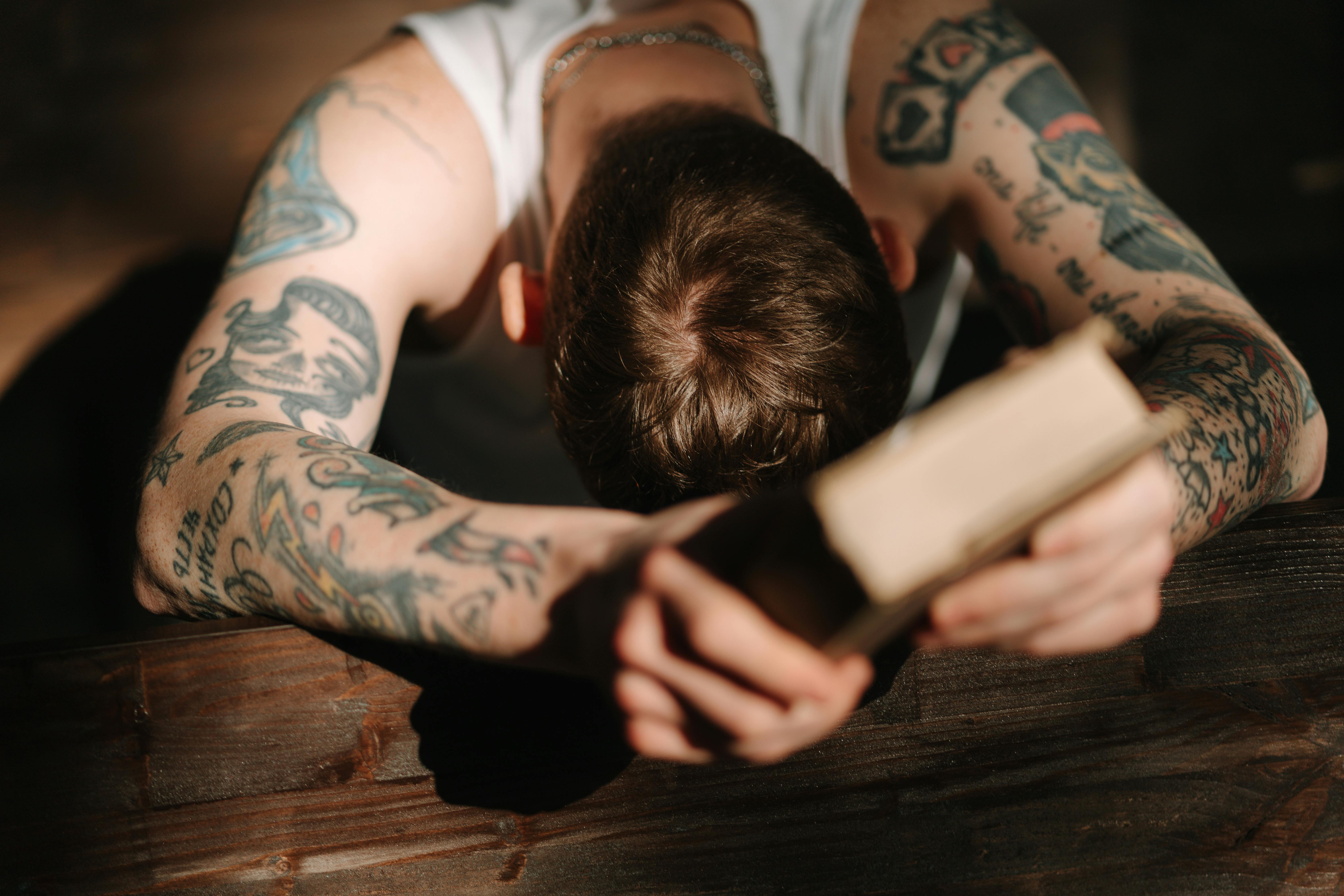 A tattooed man holding a bible bending down his head in prayer while sitting